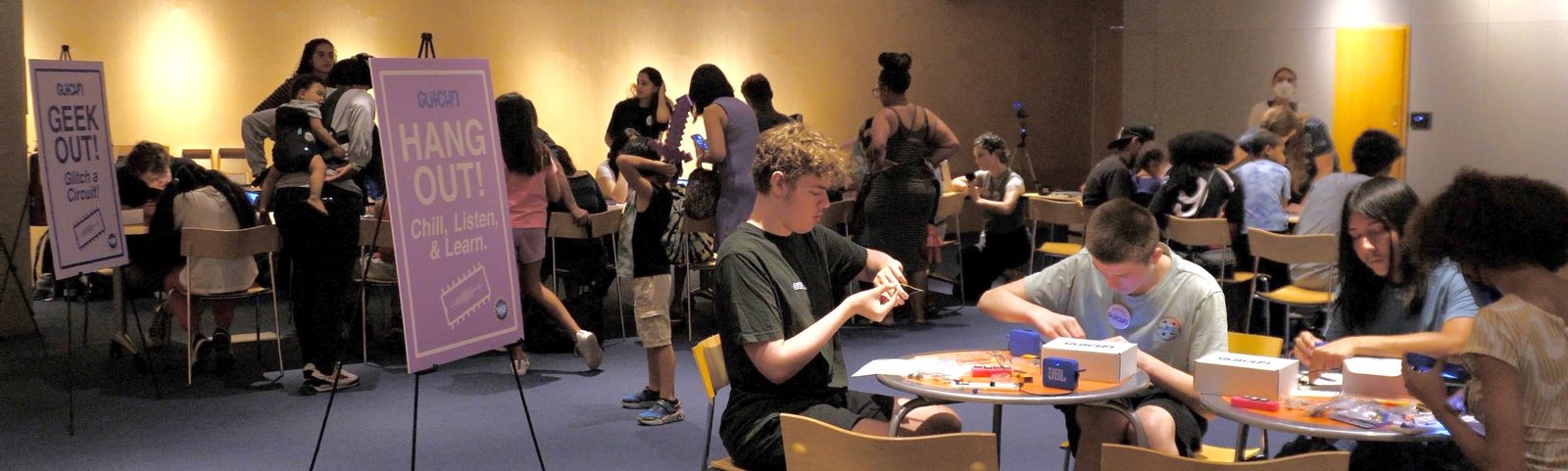 The library auditorium full of visitors building circuits at round tables, with the Hang out and Geek out signs on easels